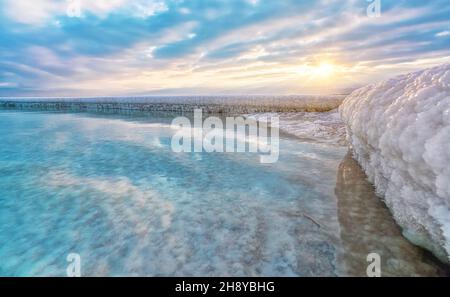 Sand komplett mit kristallinem Salz bedeckt sieht aus wie Eis oder Schnee am Ufer des Toten Meeres, türkisblaues Wasser in der Nähe, Himmel gefärbt mit Morgensonne DIS Stockfoto