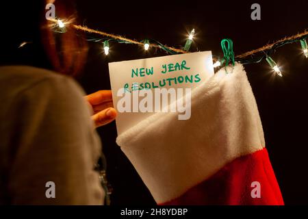 Eine Frau hängt eine Nachricht, die über die Vorsätze für ein neues Jahr zu einem Geschenkstrumpf auf einer Reihe von dekorativen Lichtern sagt. Ein skurriles, düsteres Humor-Konzept für ein Stockfoto