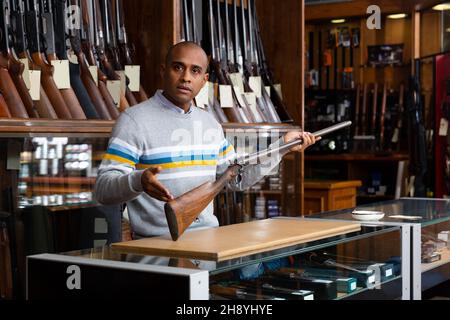 Gutaussehender, positiver erwachsener Mann im Jagdgeschäft mit seltenem Gewehr in den Händen Stockfoto