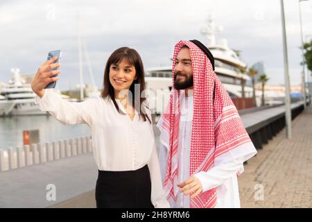 Lächelnd machten verschiedene Geschäftspartner nach einem erfolgreichen Deal Selfie im Hafen, eine junge europäische Geschäftsfrau, die während des Treffens in der Marina mit einem arabischen Kollegen ein Foto auf dem Smartphone machte Stockfoto