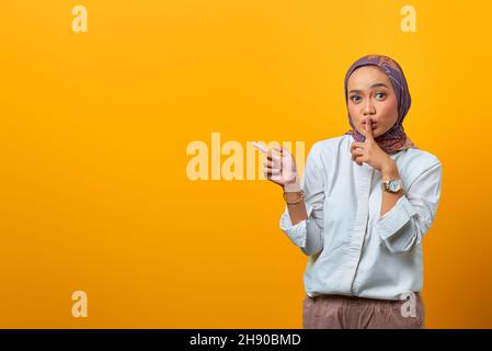 Schöne asiatische Frau Signal zur Stille und zeigt auf die Seite über gelben Hintergrund Stockfoto