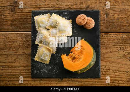 Frische, handgemachte, ungekochte italienische Ravioli-Pasta mit einer Scheibe rohem Kürbis und Makronen-Keksen auf einem kleinen Brett über einem Holztisch mit Platz zum Nachlegen Stockfoto
