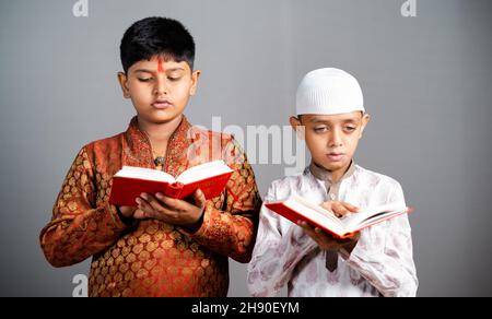 Hinduistisch-muslimische Kinder, die religiöse Schriften in traditioneller Kleidung auf grauem Hintergrund lesen - Konzept des Glaubens, des Glaubens, des Gebets zu gott und der Educatio Stockfoto