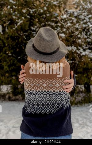 Blonde junge Frau in grauem Hut und Pullover steht mit dem Rücken. Hintergrund von weihnachtsbäumen im Schnee. Wintersaison. Stockfoto