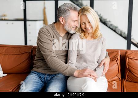 Das liebenswerte Paar mittleren Alters, verliebt, sitzt umarmt auf dem bequemen Sofa zu Hause und sieht sich zärtlich an. Konzept der Unterstützung und Fürsorge in der Ehe Stockfoto
