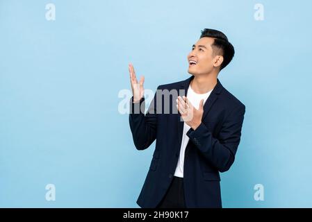 Junger lächelnder asiatischer Mann, der die Hände öffnet und seitwärts in einem isolierten hellblauen Hintergrund blickt Stockfoto