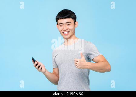 Positive glücklich lächelnden asiatischen Mann gibt Daumen nach oben, während das Smartphone in isolierten Studio blauen Hintergrund halten Stockfoto