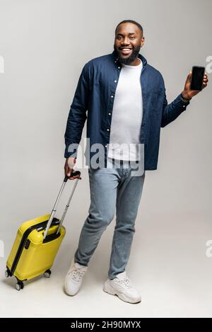 Glücklicher afrikanischer Mann, der mit einer stilvollen kleinen Handtasche und einem Handy in den Händen steht, das Telefon mit leerem Bildschirm zeigt, ein Online-Ticket hat, mit einem Komfortkonzept reist, eine vertikale Aufnahme macht Stockfoto