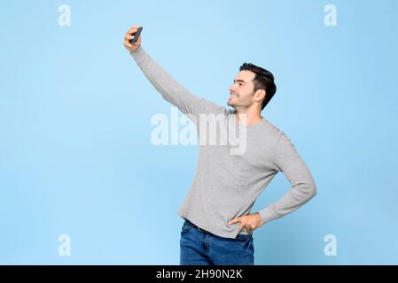 Lächelnder junger, gutaussehender Mann, der Selfie auf dem Mobiltelefon in einem isolierten hellblauen Studiohintergrund aufnahm Stockfoto