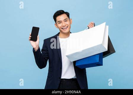 Junge gut aussehende Südost asiatischen Mann hält Handy und Einkaufen Taschen in hellblau Studio isoliert Hintergrund Stockfoto