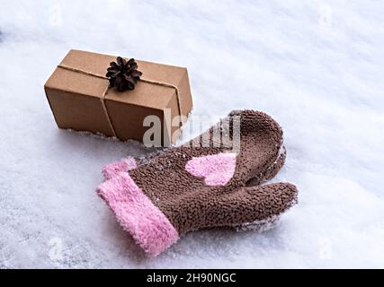 Geschenk in einer Bastelkiste mit Kieferkegel auf weißem Schnee und Fäustlinge mit rosa Herz, umweltfreundliche Öko-Konzept Frohe Weihnachten und Neujahr dekoriert Stockfoto