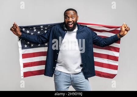 Lächelnd, schwarzer bärtiger Mann, der die amerikanische Flagge der offenen usa hinter sich hält, in lässigem Hemd und Jeans, drinnen auf grauem Hintergrund stehend, zeigt seine Unabhängigkeit und die Rechte einer gleichberechtigten Person Stockfoto
