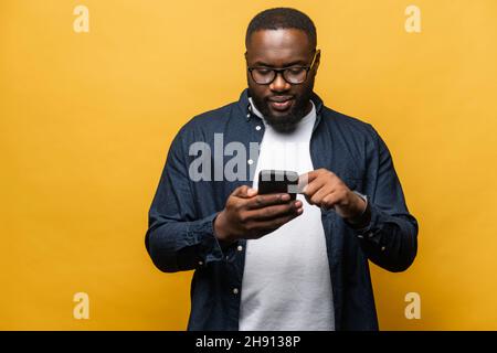 Foto von attraktiven bärtigen schwarzen Mann in Casual Shirt mit Smartphone isoliert auf grauem Hintergrund, berührt Telefonbildschirm, notwendige Werkzeug für die Produktivität, findet Inspiration online Stockfoto