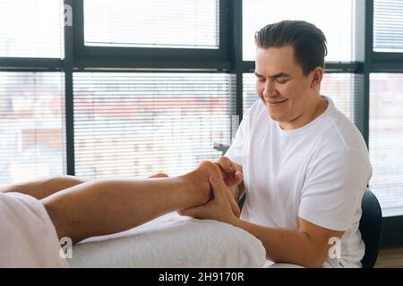 Nahaufnahme eines fröhlichen professionellen männlichen Masseurs mit starken Händen, die nach dem Sport-Training die Fersen des muskulösen Sportlers massieren. Stockfoto