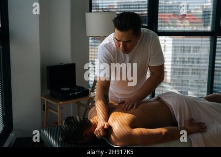 Mittlere Aufnahme eines professionellen männlichen Masseurs mit stark tätowierten Händen, die Hals und Schultern eines muskulösen Sportmanns massieren, der auf dem Bauch liegt, Stockfoto