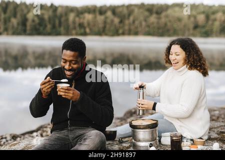 Lächelnder Mann, der mit seiner Freundin zusammen Essen isst, während er am Seeufer kampelt Stockfoto