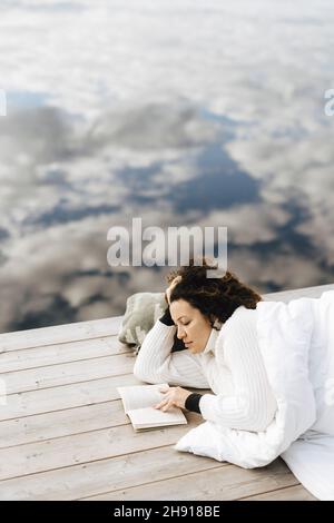 Junge Frau liest Buch, während sie auf dem Pier liegt Stockfoto