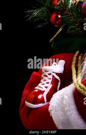 Aufnahme eines roten Weihnachtsschuhs auf einem Weihnachtsmann-Arm und Weihnachtsdekorationen im Hintergrund.das Foto wird mit künstlichem Licht auf einem Blac beleuchtet Stockfoto