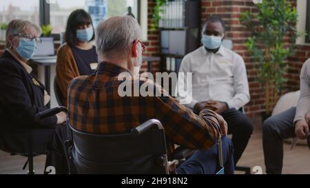 Leitender Mann mit Behinderung nimmt an einer Gruppentherapie mit Menschen während einer Pandemie Teil. Person mit Gesichtsmaske, die im Rollstuhl sitzt und beim Treffen über Alkoholabhängigkeit spricht. Stockfoto