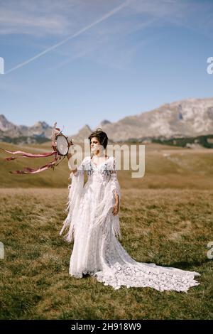 Braut in einem weißen Korbkleid mit Tamburin in den Händen steht auf einem Feld Stockfoto