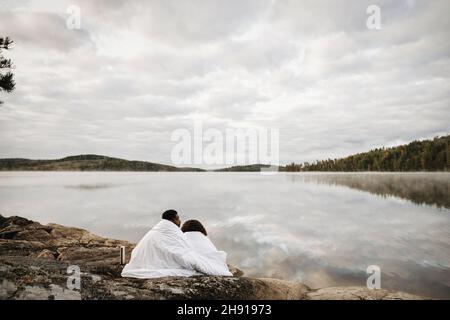 Das Paar ist in eine Decke gehüllt und sitzt am Seeufer gegen den bewölkten Himmel Stockfoto