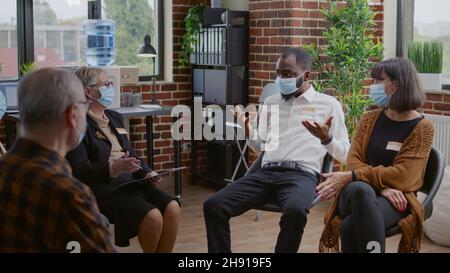 afroamerikanischer Mann spricht bei einem Therapietreffen mit einer Gruppe von Menschen über sucht während einer Pandemie. Person mit Gesichtsmaske, die psychische Probleme mit Patienten und Psychotherapeuten teilt. Stockfoto