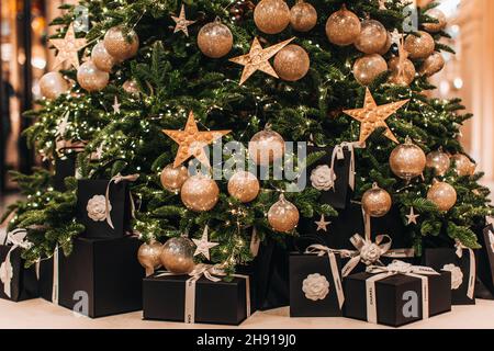 Festliches Geschenk schwarze Schachteln mit weißen Chanel-Bändern unter dem Weihnachtsbaum mit goldenen Sternen und Kugeln Stockfoto