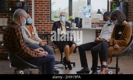 Psychotherapeut im Gespräch mit einer Gruppe von Menschen bei einem Therapietreffen über die Genesung während einer Pandemie. Frau Therapeutin trägt Gesichtsmaske, hilft Reha-Patienten mit Anleitung und Beratung Stockfoto