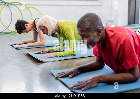 Ältere Frau trainiert auf einer Fitnessmatte in der Nähe von interrassischen Leuten, die im Fitnessstudio Plank machen Stockfoto