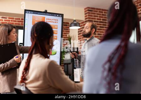 Geschäftsmann, der auf einen Monitor zeigt und den Geschäftsleuten bei der Präsentation des Unternehmens in einem Startup-Büro Marketingstatistiken erklärt. Verschiedene Geschäftsleute, die eine Geschäftspräsentation planen Stockfoto