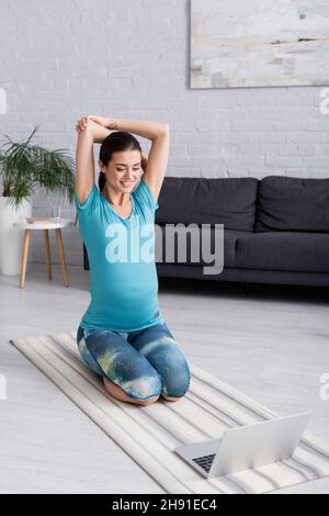 Tätowierte Schwangere Sportlerin in Sportswear beim Trainen, während sie in der Nähe des Laptops lächelte Stockfoto