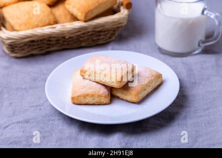 Kekse mit Puderzucker auf einem weißen Teller. Im Hintergrund ein Korb mit Keksen und eine Tasse Milch. Nahaufnahme. Stockfoto