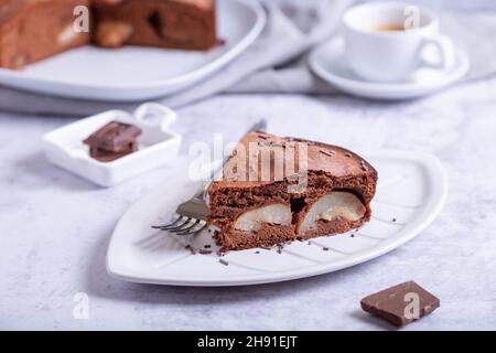 Stück Schokoladenkuchen mit Birne. Im Hintergrund ist eine Torte, eine Tasse Kaffee und Schokoladenstücke. Nahaufnahme. Stockfoto
