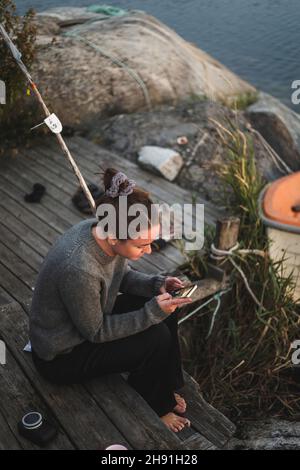Frau, die im Urlaub auf dem Pier sitzt und ein Smartphone benutzt Stockfoto