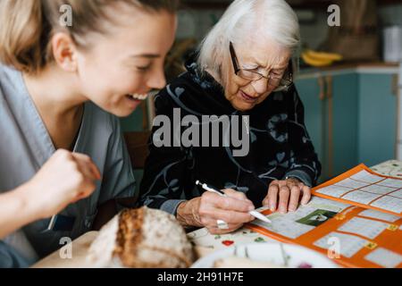 Lächelnde weibliche Betreuerin schaut, während ältere Frau Kreuzworträtsel in der Küche löst Stockfoto