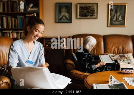 Glückliche weibliche Pflegekraft Faltdecke während ältere Frau Zeitung zu Hause lesen Stockfoto