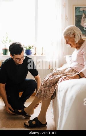 Lächelnde ältere Frau schaut, während ein Krankenschwester ihr hilft, Sandalen ins Schlafzimmer zu legen Stockfoto