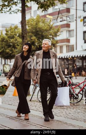 Ein älteres Paar hält sich die Hände, während es in der Stadt auf einem Fußweg läuft Stockfoto