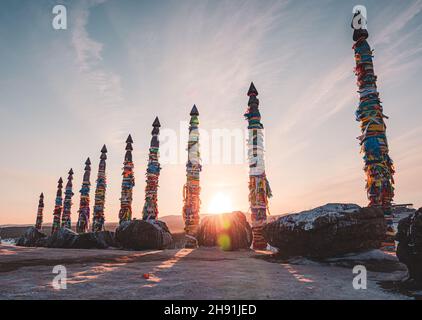 Traditionelle buryat Schamanen-Säulen mit bunten Bändern im Winter bei Sonnenuntergang, Kap Burkhan, Olkhon Insel. Winter Baikal. Stockfoto