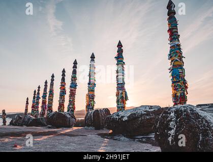 Traditionelle buryat Schamanen-Säulen mit bunten Bändern im Winter bei Sonnenuntergang, Kap Burkhan, Olkhon Insel. Winter Baikal. Stockfoto