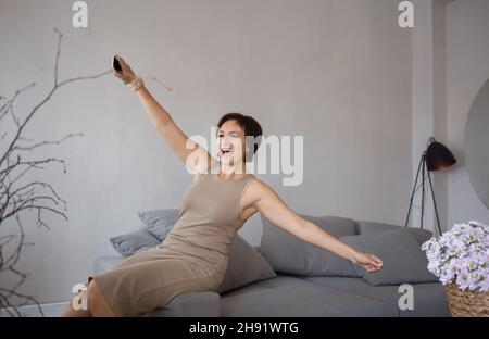 Sehr zufrieden schöne junge Frau in einem engen Kleid glücklich spreizte ihre Arme sitzt auf der Couch. Freudig elegante Geschäftsfrau feiert Triumph, hap Stockfoto