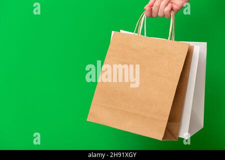 Weibliche Hand hält zwei Einkaufstaschen isoliert auf grünem Hintergrund. Weiß und braun basteln leere Papiertüten in der Hand. Black friday Verkauf, Rabatt, Shopping und Ökologie Konzept Stockfoto