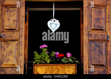 Offenes Holzfenster mit Stottern, die mit Blumen im Blumentopf dekoriert sind, und weiße herzförmige Dekoration, die von oben hängt - ländliches alpenhaus im Stil Stockfoto