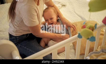 Die junge Mutter wechselt die Clithes und kleidet ihren kleinen Sohn nachts im Bett. Konzept der Elternschaft, Elternermüdung und Kinderentwicklung. Stockfoto