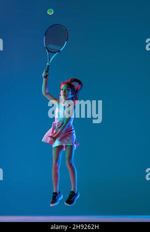 Porträt eines kleinen Mädchens, Tennisanfänger, der Tennis spielt, isoliert auf blauem Studiohintergrund in Neonlicht. Sport, Studium, Kinderkonzept Stockfoto