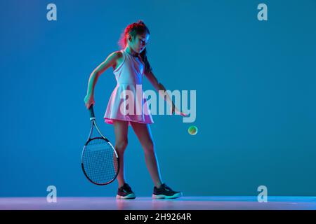 Ein kleines Mädchen, Anfänger Tennisspielerin posiert isoliert auf blauem Studiohintergrund in Neonlicht. Sport, Studium, Kinderkonzept Stockfoto