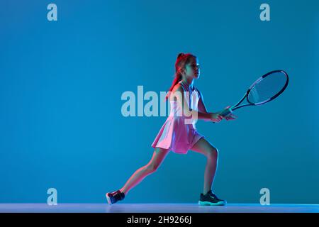 Porträt eines kleinen Mädchens, Tennisanfänger, der Tennis spielt, isoliert auf blauem Studiohintergrund in Neonlicht. Sport, Studium, Kinderkonzept Stockfoto