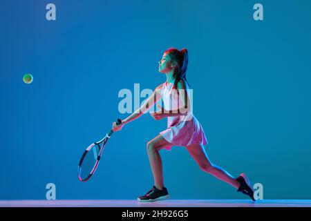 Porträt eines kleinen Mädchens, Tennisanfänger, der Tennis spielt, isoliert auf blauem Studiohintergrund in Neonlicht. Sport, Studium, Kinderkonzept Stockfoto