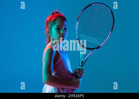 Halblanges Porträt eines kleinen Mädchens, eines Tennisanfängers, der mit einem Schläger posiert, der auf blauem Studiohintergrund in Neonlicht isoliert ist. Sport, Studium Stockfoto