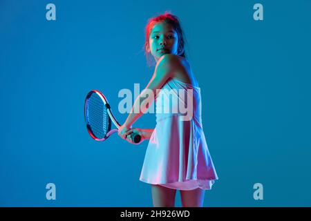 Porträt eines kleinen Mädchens, Tennisanfänger, der Tennis spielt, isoliert auf blauem Studiohintergrund in Neonlicht. Sport, Studium, Kinderkonzept Stockfoto